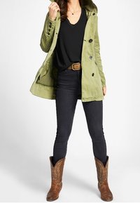 Olivgrön huoodjacka med knäppningar, matchad med en svart topp, mörka jeans och bruna cowboyboots med dekorativt stygn.