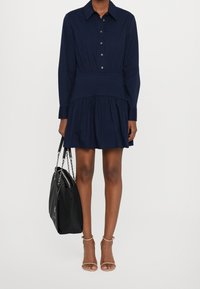 Robe bleu marine à boutons, avec une jupe plissée, des manches longues et une taille ajustée. Associée à un sac à main noir et des talons ouverts nude.