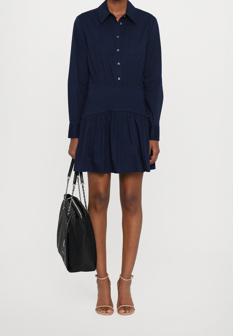 Robe bleu marine à boutons, avec une jupe plissée, des manches longues et une taille ajustée. Associée à un sac à main noir et des talons ouverts nude.