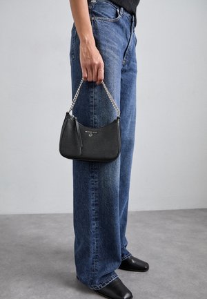 Persoon draagt blauwe spijkerbroek en zwarte schoenen en houdt een kleine zwarte handtas met een zilveren kettinghengsel naast zich.