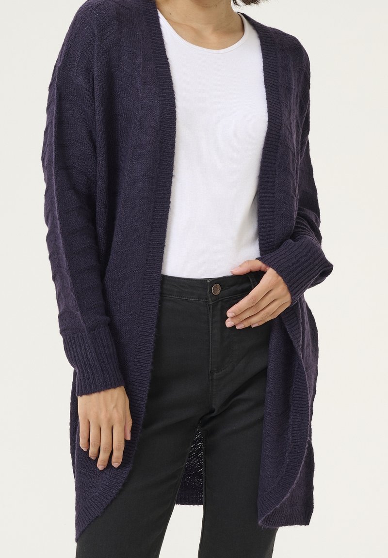 Personne portant un cardigan en tricot violet foncé par-dessus une chemise blanche et un pantalon noir, avec une main dans la poche et l'autre détendue.