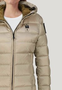 Giacca puffer beige con zip frontale, cappuccio e tasche laterali. Presenta una texture trapuntata e una fodera interna a contrasto. Logo sul petto.