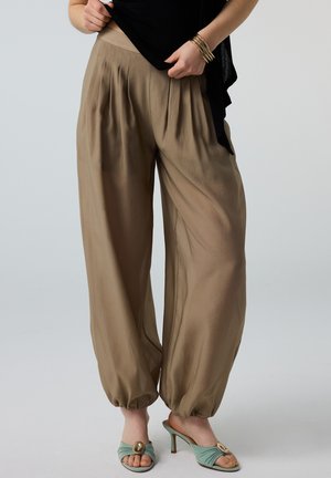 Femme portant un pantalon ample beige taille haute avec des poignets élastiques, un top noir, des bracelets dorés et des sandales à talons ouvertes vertes.