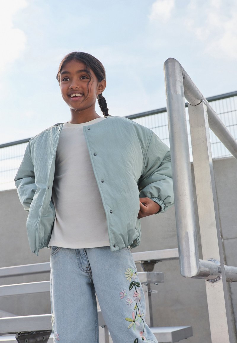 Ung flicka ler utomhus, bär en ljusgrön jacka och blå jeans med blommig brodyr, står nära metallräcke och läktare.