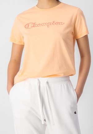 Personne portant un t-shirt Champion pêche et un pantalon de survêtement blanc avec cordon de serrage, les mains dans les poches, cadrée au niveau du cou et des genoux.