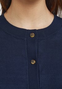 Detailní záběr na tmavě modrý pletený kardigan se dvěma hnědými knoflíky zapnutými u límce na osobě s hnědými vlasy.