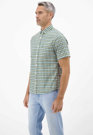 Mand iført kortærmet grøn og beige ternet skjorte med lyseblå jeans, stående mod en ensfarvet hvid baggrund.