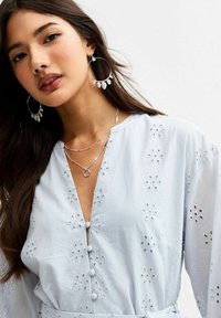 Lichtblauwe blouse met knoopsluiting, voorzien van bloemembroidery, witte knopen en een v-halslijn, gecombineerd met zilveren sieraden en creolen.