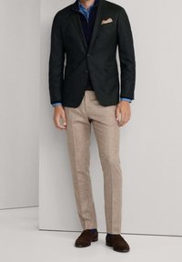 Un homme porte un blazer noir, un pull marine, une chemise bleu clair, un pantalon beige et des mocassins en daim marron, debout contre un mur clair uni.