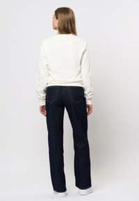 Vrouw met lang bruin haar staat met haar rug naar ons gekeerd, draagt een wit langslobige top, donkerblauwe jeans en witte sneakers tegen een effen achtergrond.