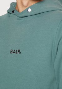 Gros plan sur un sweat à capuche bleu sarcelle avec un détail métallique hexagonal argenté sur la capuche et le logo noir "BALR." sur la poitrine.