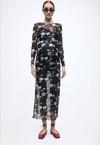 Μαύρο διαφανές φόρεμα με λουλουδένια σχέδια, μακριά μανίκια και εφαρμοστή σιλουέτα. Συνδυάζεται με κόκκινα πλατφόρμα παπούτσια και μοντέρνα γυαλιά ηλίου.