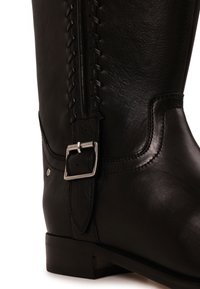 Gros plan d'une botte en cuir noir avec des coutures décoratives et une boucle carrée argentée sur la bride latérale.