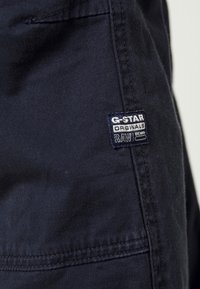 Marinblå G-Star RAW denim med en sydd etikett med logotyp, subtil textur och en närbild av tyget och sömmarna.