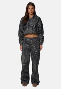 Korte hoodie en wijde broek in een donkere camouflageprint, met een zachte textuur en een trekkoord in de taille voor comfort. Bruinachtig schoeisel maakt de look compleet.