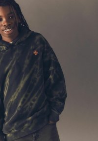 Schwarzer und grüner Batik-Hoodie mit einem orangen Totenkopf-Logo auf der linken Brust. Der Stoff wirkt weich, hat eine lockere Passform und eine Kängurutasche.