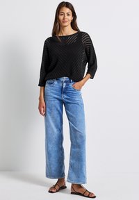 Schwarzer Strickpullover mit Zickzackmuster und weiten Ärmeln, kombiniert mit hellblauen weit geschnittenen Jeans und flachen schwarzen Sandalen.