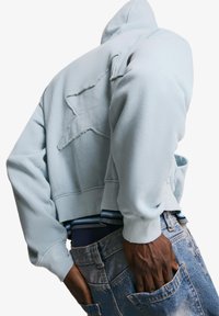 Sweatshirt court bleu clair avec un design de star texturé au dos et des poignets côtelés, associé à un jean bleu déchiré.