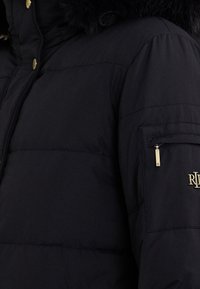 Černá péřová bunda s texturovaným povrchem, vybavená odnímatelnou kožešinovou kapucí, zlatými kovovými prvky a kapsou na zip na rukávu.