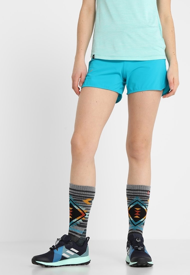 Personne portant un short bleu clair, des chaussettes hautes à motifs géométriques, et des chaussures de sport noires et bleues, debout sur un fond blanc.