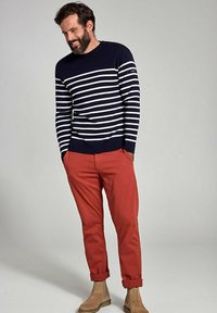 Maglione a righe blu marino e bianco con scollatura rotonda, polsini blu marino e quattro bottoni sulle spalle, abbinato a pantaloni rossi arrotolati e stivali beige.