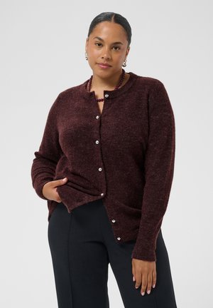Femme aux cheveux foncés portant un cardigan bordeaux boutonné, un pantalon noir et un collier de perles, debout avec une main dans sa poche.