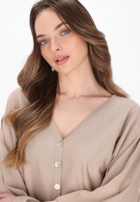 Béžový cardigan s výstřihem do V vyrobený z měkké pleteniny, s pěti perlami podobnými knoflíky, dlouhými rukávy a uvolněným střihem.