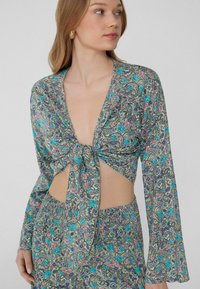 Kwiatowy crop top z lekkiego materiału z wiązanym przodem, głębokim dekoltem w serek i długimi rękawami, z kolorowym wzorem paisley w turkusie i różu.