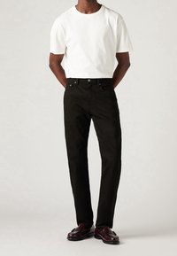 Mann trägt ein schlichtes weißes T-Shirt mit kurzen Ärmeln, schwarze Jeans mit geradem Bein und dunkelburgunderrote Loafers, steht mit den Händen hinter dem Rücken.