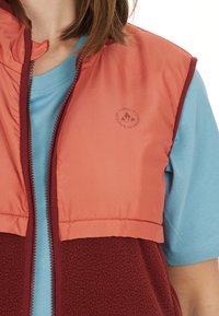 Fleeceväst med röd kropp, lätta orange axlar och en blå T-shirt under. Har en logo på bröstet och en dragkedja.