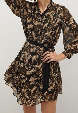 Robe noire transparente imprimée paisley doré, manches longues bouffantes, devant boutonné et ceinture noire nouée à la taille.