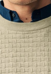 Pull en tricot beige clair avec un motif de tissage en panier texturé, comprenant une chemise col bleu marine en dessous avec des détails de boutons.