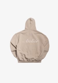 Sudadera de color beige claro con capucha ajustable, puños y cinturilla acanalados. Presenta un bordado de "Atelier" en la parte posterior. Textura de tela suave.