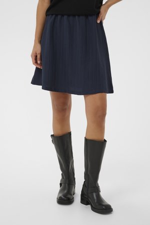 Granatowa plisowana spódnica noszona z czarnymi skórzanymi botkami do połowy łydki na osobie stojącej na jednolitym tle.