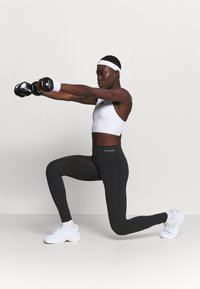 Vrouw in sportkleding maakt een uitvalspas naar voren terwijl ze twee dumbbells van 10 kg recht voor zich houdt tegen een effen achtergrond.