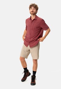 Camisa de cuadros roja de manga corta con un bolsillo frontal, combinada con shorts beige y botas de senderismo marrones. Medias negras a rayas visibles.