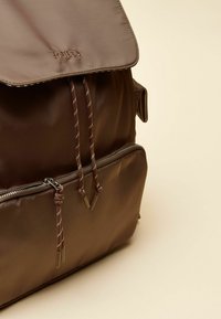 Rjavi nahrbtnik iz najlona z zložljivim preklopom, zateznim vrvnim zapiranjem in sprednjim žepom z zadrgo. Ima kovinske poudarke in kontrastno šivanje.