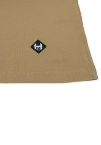 Tan bomuld t-shirt med en glat tekstur, som har en sort diamanformet logo-patch med et "R" i nederste venstre hjørne.