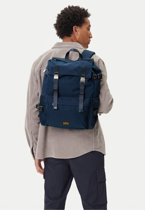 Uomo con camicia di velluto beige e pantaloni scuri che porta uno zaino blu navy con fibbie argentate, visto da dietro.