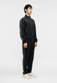 Μαύρη φόρμα με λευκές ρίγες στα μανίκια και τα πόδια. Διαθέτει ζακέτα με φερμουάρ και άνετη εφαρμογή. Συνδυάζεται με ανοιχτόχρωμα αθλητικά παπούτσια.