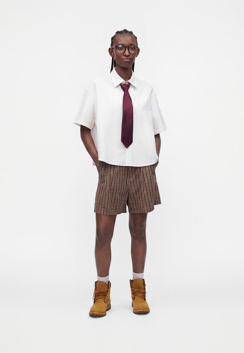 Person with glasses wearing a white short-sleeve shirt, burgundy tie, plaid shorts, white socks, and brown lace-up boots standing with hands in pockets.