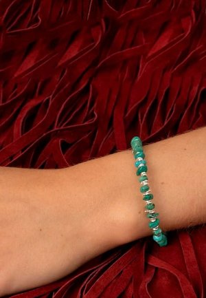 Bras reposant sur un tissu rouge texturé, portant un bracelet turquoise en perles avec de petits accents argentés.