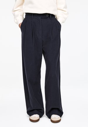 Navy pleated trousers made from a textured fabric, featuring side pockets and a button closure, paired with light-colored sneakers.