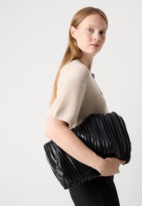 Žena s dlhými blond vlasmi má na sebe béžový pletený top a drží veľkú čiernu skladanú kabelku pred bielym pozadím.