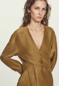 Abito di colore bronzo, con texture, scollatura profonda a V e maniche lunghe, dotato di un punto vita legato e una leggera lucentezza. Accentuato da una collana con pendente.