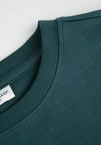 Ciemnoturkusowa bluza z prążkowanym okrągłym dekoltem, gładką fakturą oraz metką widoczną na wewnętrznej stronie kołnierza. Prosty design bez wzorów.