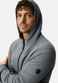 Grijze hoodie met rits en een zachte textuur, voorzien van een zwarte ritssluiting aan de voorkant, een capuchon en een zwart logoplaatje op de linker mouw.