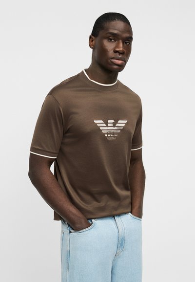 Καφέ μπλουζάκι με αντίθετο λευκό γιακά και τελειώματα στα μανίκια, με οριζόντιες ρίγες σε λογότυπο στο κέντρο. Κανονική εφαρμογή.