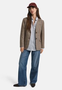 Donna che indossa un blazer a quadri marrone, una camicia button-up azzurra chiara, jeans a gamba larga, scarpe scure e un cappellino da baseball bordeaux, in piedi contro uno sfondo bianco.