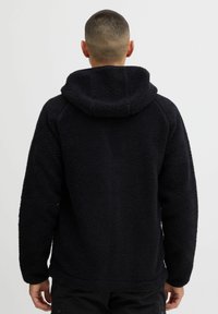 Sudadera de forro polar negra con una superficie texturizada y capucha, con mangas raglán y un corte holgado, vista desde la parte de atrás.